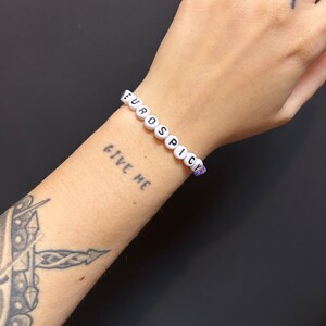May include: White beaded bracelet with the words "EUROSPICY" spelled out in black letters. The bracelet is worn on a person's wrist with a tattoo of the words "GIVE ME" on their arm.