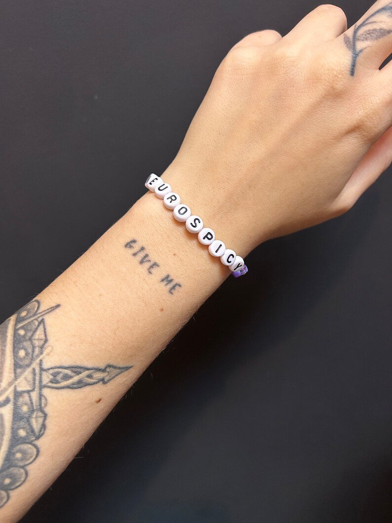 May include: White beaded bracelet with the words "EUROSPICY" spelled out in black letters. The bracelet is worn on a person's wrist with a tattoo of the words "GIVE ME" on their arm.