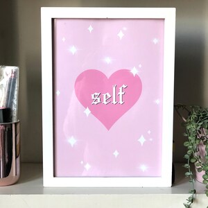 May include: A white framed print with a pink background and a pink heart with the word "self" in white gothic lettering. The background is decorated with white stars.