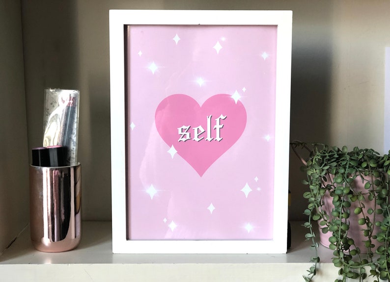 May include: A white framed print with a pink background and a pink heart with the word "self" in white gothic lettering. The background is decorated with white stars.