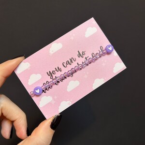 May include: A purple beaded bracelet with white heart charms. The bracelet is on a pink card with white clouds and stars. The card says "You can do all things but fail."