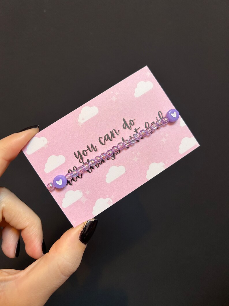 May include: A purple beaded bracelet with white heart charms. The bracelet is on a pink card with white clouds and stars. The card says "You can do all things but fail."