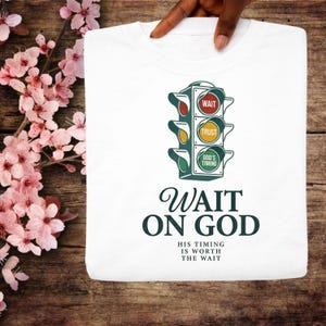 Puede incluir: Camiseta blanca con un diseño de semáforo que muestra las palabras «WAIT», «TRUST» y «GOD'S TIMING». Debajo del gráfico, las palabras «WAIT ON GOD» se muestran en una fuente elegante, con la frase «HIS TIMING IS WORTH THE WAIT» debajo.