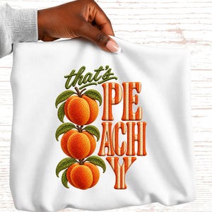 May include: White sweatshirt with a graphic design featuring three orange peaches with green leaves and the text "that's PEACHY" in orange and white. The design has a vintage, retro style.