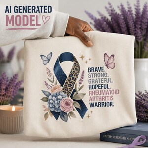 May include: A cream-colored sweatshirt featuring a blue awareness ribbon with a leopard print section, surrounded by floral illustrations and butterflies. The text reads: BRAVE. STRONG. GRATEFUL. HOPEFUL. RHEUMATOID ARTHRITIS WARRIOR.