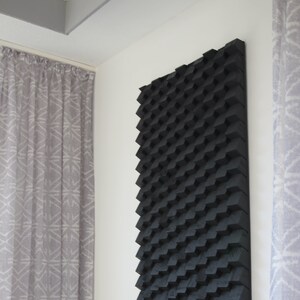 Puede incluir: Panel de pared negro y geométrico con un diseño texturizado tridimensional. El panel es rectangular y está montado en una pared blanca. El diseño está hecho de pequeños bloques angulados. Cortinas visibles a ambos lados.