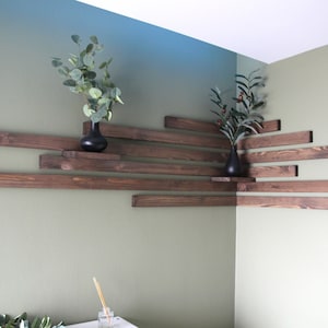 Può includere: Un set di mensole in legno marrone scuro montate in un angolo di una stanza. Le mensole sono disposte a scalare e sono decorate con due piccole piante in vaso.