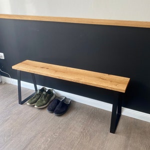 Op de afbeelding: Een houten bank met zwarte metalen poten. De bank staat voor een zwarte muur met een witte plint. Er liggen twee paar schoenen op de vloer voor de bank.