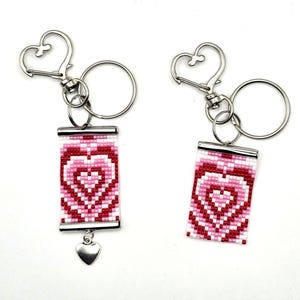 May include: Two keychains with heart-shaped designs. Each keychain features a rectangular beaded pendant with a pink and red heart pattern. The keychains have silver-colored hardware, including a heart-shaped clasp and a small heart charm.