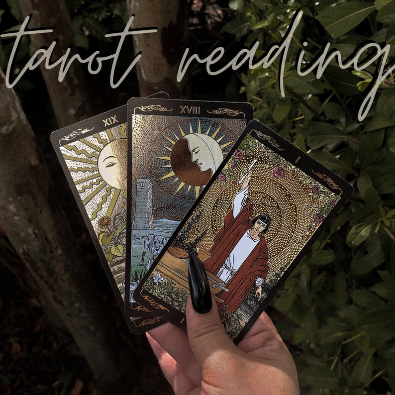 May include: Three tarot cards with gold accents and black borders. The cards are numbered XIX, XVIII, and a card with a sword. The cards are held in a hand with black nail polish.