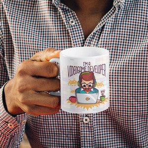 Puede incluir: Taza de cerámica blanca con una ilustración de dibujos animados de una persona con auriculares y gafas, sentada frente a un ordenador. El texto "I'm a Videogame Developer" está impreso en la taza.
