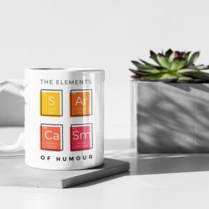 May include: White ceramic mug with a black outline and the text "The Elements of Humour" printed on the front. The mug features four colorful squares with the chemical symbols "S", "Ar", "Ca", and "Sm" inside.