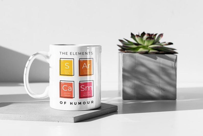 May include: White ceramic mug with a black outline and the text "The Elements of Humour" printed on the front. The mug features four colorful squares with the chemical symbols "S", "Ar", "Ca", and "Sm" inside.
