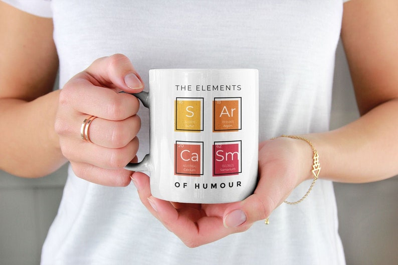 May include: White ceramic mug with the text "The Elements of Humour" and four chemical element boxes with the symbols "S", "Ar", "Ca", and "Sm".