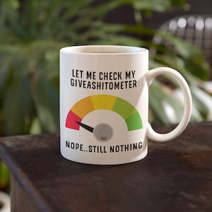Könnte beinhalten: Weiße Keramiktasse mit einer schwarzen und roten Grafik einer Anzeige, die "Let me check my giveashitometer" und "Nope...still nothing" anzeigt.