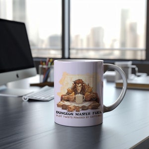 May include: White ceramic mug with a fantasy illustration of a character and the text "DUNGEON MASTER FUEL PLOT TWISTS POWERED BY CAFFEINE!". The mug has a white handle and is set on a dark surface.