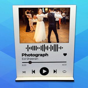 Op de afbeelding: Een wit houten fotolijstje met een zwart-wit foto van een dansend stel. Het lijstje heeft een zwarte Spotify-achtige muziekspelerinterface met het nummer "Photograph" van Ed Sheeran dat wordt afgespeeld.