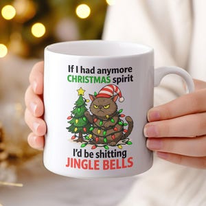 May include: White ceramic mug with a humorous Christmas design. The mug features a grumpy cartoon cat wearing a Santa hat and wrapped in Christmas lights, with the text "If I had anymore CHRISTMAS spirit I'd be shitting JINGLE BELLS."