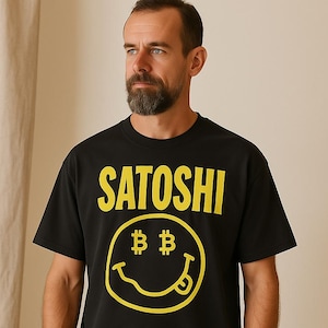 Puede incluir: Camiseta negra con la palabra "SATOSHI" en letras amarillas en bloque sobre una carita sonriente amarilla con símbolos de Bitcoin para los ojos y una lengua fuera. La camiseta es de cuello redondo y manga corta.