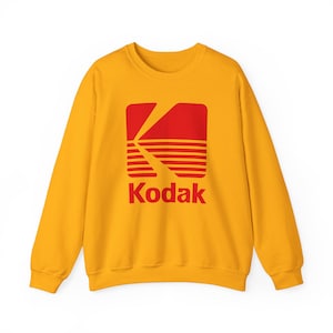 Puede incluir: Sudadera amarilla mostaza con el logotipo de Kodak en rojo. El logotipo incluye una forma de 'K' estilizada y líneas horizontales, con el nombre de la marca debajo. Cuello redondo clásico.