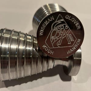 May include: Close-up of three brushed metal objects. Two are cylindrical with ridged sides, and the third is a circular coin with the text "WISEMAN KOCH GLOVER HANSEN" around a central design. The objects are on a white surface.