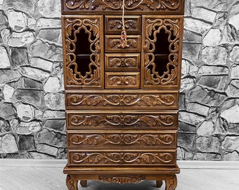 May include: A tall, intricately carved wooden cabinet with six drawers and two glass doors. The cabinet has a dark brown finish and ornate details.