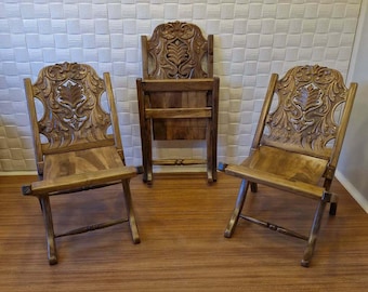 May include: Three wooden folding chairs with intricate carved designs on the backrests. The chairs are made of dark brown wood and have a rustic look.