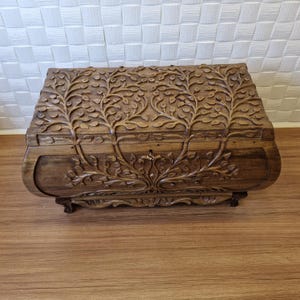 Puede incluir: Un cofre de madera hecho a mano con intrincadas tallas de árboles y hojas. El cofre es de color marrón cálido y tiene un diseño curvo. La tapa y la parte delantera presentan tallas en relieve detalladas de árboles y follaje.