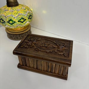 May include: A dark brown wooden puzzle box with a carved floral design on the lid. The box has a rectangular shape with vertical wooden slats around the sides. The box is sitting on a white surface.