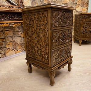 May include: A brown wooden nightstand with three drawers, intricately carved with floral and scroll patterns. The nightstand has a rectangular shape and stands on four carved legs. A second nightstand is visible in the background.