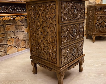 May include: A brown wooden nightstand with three drawers, intricately carved with floral and scroll patterns. The nightstand has a rectangular shape and stands on four carved legs. A second nightstand is visible in the background.