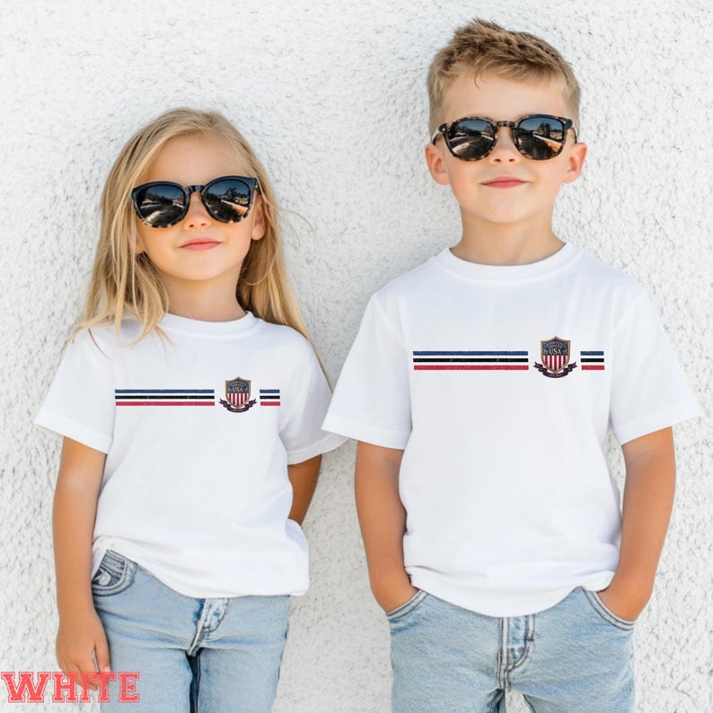 May include: Two white short-sleeve t-shirts featuring a horizontal stripe pattern in navy, white, and red across the chest. Centered on the stripes is a shield-shaped emblem with the text USA. The shirts are worn with blue denim jeans.