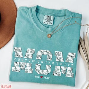May include: Seafoam green t-shirt with the word "MOM" in white floral print letters. Below the word "MOM" is the text "COSTUM UNIVERSITY" and "EST. YEAR" with a red heart. A gold necklace with a pendant is on the shirt.