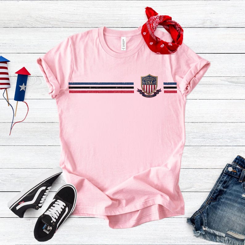 May include: A light pink short-sleeve t-shirt featuring a horizontal stripe pattern in navy, white, and red across the chest. A United States Soccer crest with the text "UNITED STATES SOCCER USA" is printed on the right side of the chest stripe.