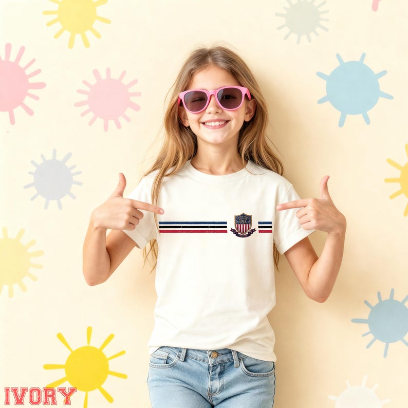 May include: A short-sleeved white t-shirt featuring a horizontal graphic design with three thin stripes in navy, white, and red, accompanied by a shield-shaped emblem containing the text USA. The shirt is paired with blue denim jeans.