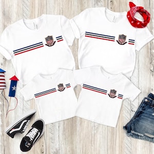 May include: Four white short-sleeve t-shirts featuring a horizontal stripe pattern in red, white, and blue. Each shirt displays a central shield emblem with the text United States USA. The set includes a red bandana, black sneakers, and denim shorts.