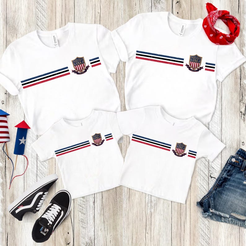 May include: Four white short-sleeve t-shirts featuring a horizontal stripe pattern in red, white, and blue. Each shirt displays a central shield emblem with the text United States USA. The set includes a red bandana, black sneakers, and denim shorts.