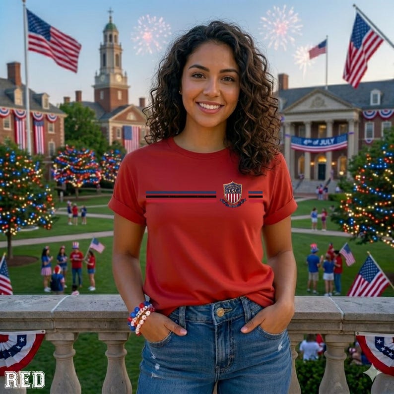May include: A red short-sleeved t-shirt featuring a horizontal dark blue stripe across the chest and a small shield-shaped emblem with the text USA in the center. The shirt is paired with blue denim jeans and a multi-colored beaded bracelet.