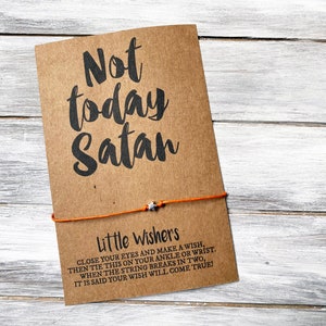 Puede incluir: Una tarjeta marrón con el texto "Not today Satan" en escritura negra. Debajo, se lee "Little Wishers" con instrucciones. Una cuerda naranja con un pequeño dije de estrella está adjunta.