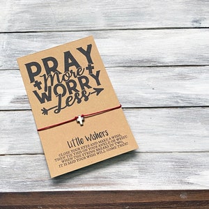 May include: A red string bracelet with a silver cross charm and a card that says "Pray More Worry Less" and instructions on how to make a wish.