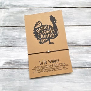 Puede incluir: Una tarjeta marrón con una ilustración de pavo negro y el texto "Happy Thanksgiving". Un cordón marrón con un dije de estrella plateada está unido a la tarjeta. La tarjeta tiene el texto "Little Wishers" e instrucciones para hacer un deseo.