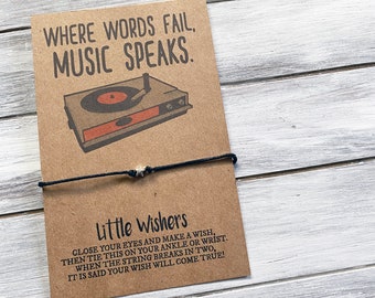 Peut inclure: Une carte marron avec le texte "WHERE WORDS FAIL, MUSIC SPEAKS." et une image de dessin animé d'un tourne-disque. Un bracelet en cordon noir avec un charme étoile argenté est attaché à la carte. La carte indique "Little Wishers CLOSE YOUR EYES AND MAKE A WISH, THEN TIE THIS ON YOUR ANKLE OR WRIST. WHEN THE STRING BREAKS IN TWO, IT IS SAID YOUR WISH WILL COME TRUE!"