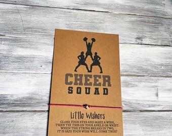 May include: A brown card with the words "CHEER SQUAD" and a cheerleading graphic. Below, the text "Little Wishers" and instructions for tying a wish bracelet. A pink string with a small star charm is attached.