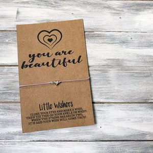 May include: A brown card with the text "you are beautiful" and a silver heart charm attached to a light purple string. The card also includes instructions for a wish bracelet: "Close your eyes and make a wish, then tie this on your ankle or wrist. When the string breaks in two, it is said your wish will come true!"