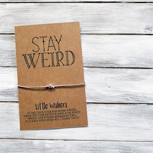 May include: A brown card with the text "STAY WEIRD" printed in black. A thin purple string is tied around the card with a small silver bead in the center. The card also has the text "Little Wishers" and instructions for making a wish.