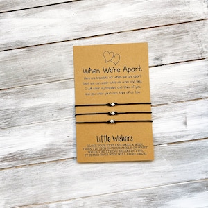 Op de afbeelding: Twee zwarte vriendschapsarmbanden met zilveren sterbedels en een kaart met de tekst "When We're Apart" en "Little Wishers". De kaart legt uit dat de armbanden zijn voor wanneer vrienden uit elkaar zijn en dat wanneer de draad breekt, een wens uitkomt.