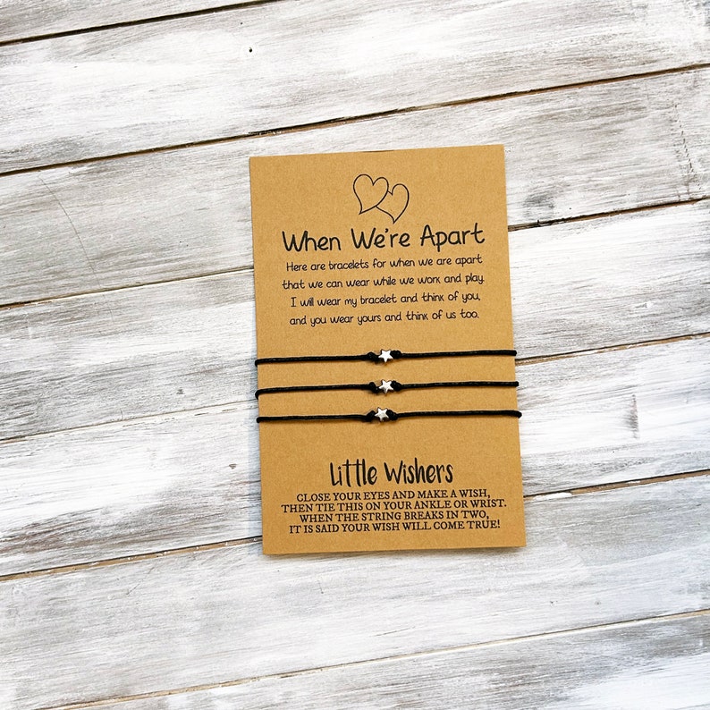 Op de afbeelding: Twee zwarte vriendschapsarmbanden met zilveren sterbedels en een kaart met de tekst "When We're Apart" en "Little Wishers". De kaart legt uit dat de armbanden zijn voor wanneer vrienden uit elkaar zijn en dat wanneer de draad breekt, een wens uitkomt.