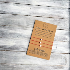 May include: Two red string bracelets with white heart charms and a card that reads "When We're Apart" and "Little Wishers". The card explains that the bracelets are for when you are apart and that you can make a wish when the string breaks.