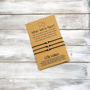 Op de afbeelding: Twee zwarte vriendschapsarmbanden met zilveren ster-bedels en een kaart met de tekst "When We're Apart" en "Little Wishers". Sluit je ogen en wens iets, bind het dan om je enkel of pols. Wanneer het touwtje in twee&euml;n breekt, wordt gezegd dat je wens uitkomt!