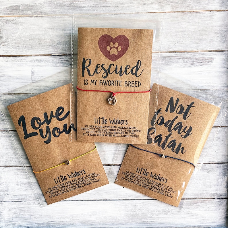 May include: Three brown cardstock tags with red, yellow, and blue string bracelets attached. Each tag has a paw print charm and a different saying: "Rescued is my favorite breed", "Love you", and "Not today Satan". Each tag includes the text "Little Wishers" and instructions for making a wish.